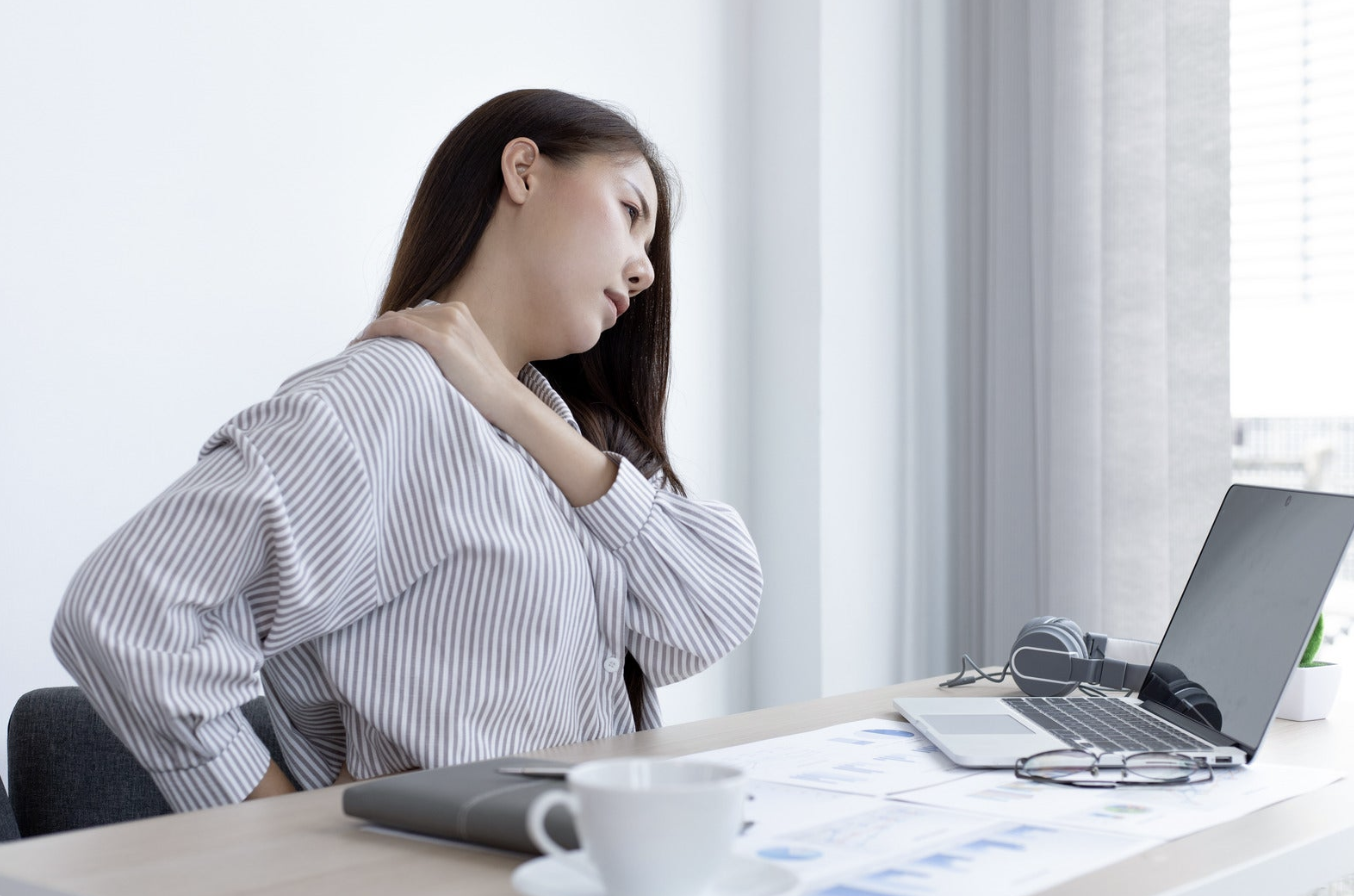 A woman at an office desk holding her stiff neck and lower back, illustrating why people wait to see a chiropractor until pain worsens.